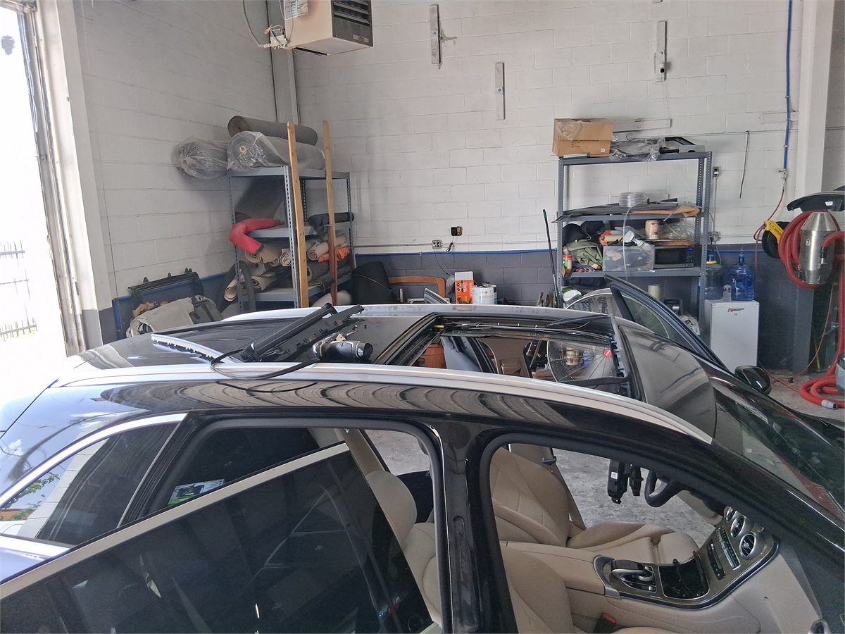 A factory sunroof glass panel lifted out and resting on the roof of a dark sedan at the Sunroof King shop in Houston, with the roof opening, frame and mechanism exposed below it